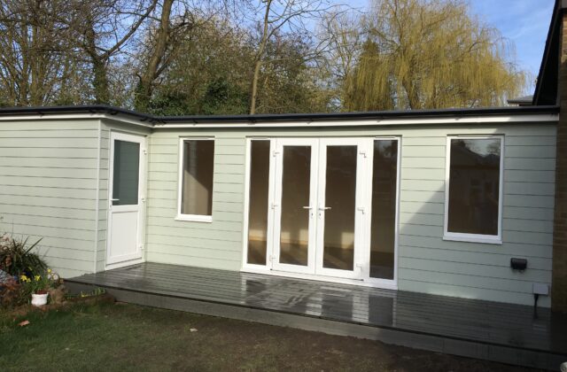 L-shaped bespoke garden room in Shelford clad in sage hardie board with french doors, twin casement windows and a raised anthracite composite decking veranda