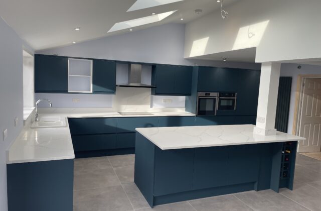 Open-plan kitchen in a single-storey lean-to extension in Cherry Hinton, Cambridge, with a vaulted ceiling, Velux skylights and dark teal handleless units
