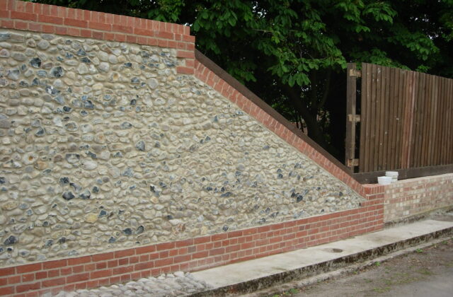 Flint wall with red brick quoins and a raked brick coping stepping down a slope