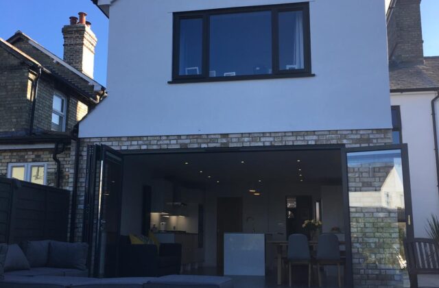Two-storey rear extension in Royston with white painted render, dark-framed glazing and full-width anthracite grey bifold doors onto a flush patio terrace