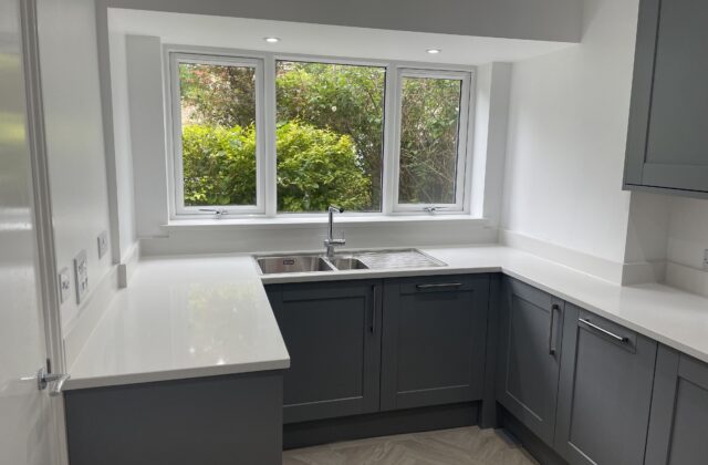 U-shaped slate-grey shaker kitchen in a bay window extension with white quartz worktops and a stainless steel sink and drainer